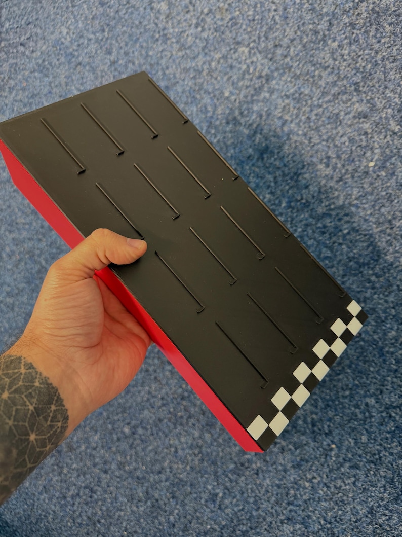 May include: A black and red rectangular box with a checkered pattern on one end. The top surface is black with evenly spaced, thin, raised lines. The box is held in a hand.