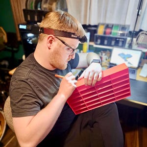 artisan crafting a handmade 3D-printed car display shelf in a home workshop. The maker carefully refines details, ensuring quality and durability. Behind him, spools of filament and a design on screen highlight the custom, small production process