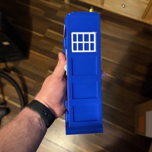 May include: A blue toy police box with a white window panel. The toy has a rectangular shape with three rectangular panels on the front. The toy is held in a person's hand.