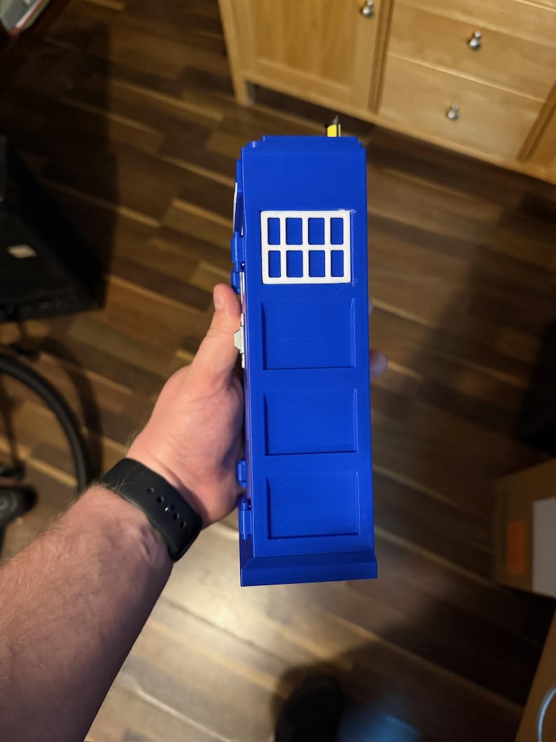 May include: A blue toy police box with a white window panel. The toy has a rectangular shape with three rectangular panels on the front. The toy is held in a person's hand.