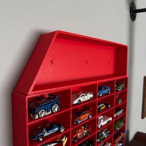 May include: Red plastic wall-mounted storage unit with a triangular roof and multiple compartments for displaying toy cars. The unit is filled with various toy cars in different colors and styles.
