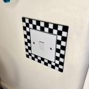 May include: A white light switch with a black and white checkered border. The switch is mounted on a white wall. The border is a square shape with alternating black and white squares.