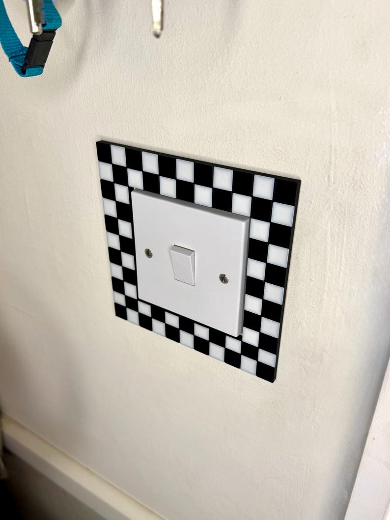 May include: A white light switch with a black and white checkered border. The switch is mounted on a white wall. The border is a square shape with alternating black and white squares.