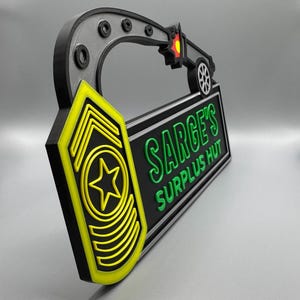 May include: A 3D-printed sign with the text "SARGE'S SURPLUS HUT" in green on a black background. The sign has a yellow and black military insignia and a black and gray handle with a small vehicle design. The sign is approximately 12 inches long.