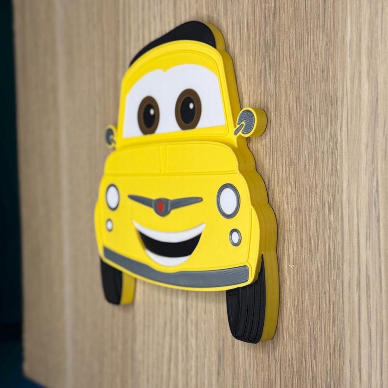 May include: A yellow car-shaped wall decoration featuring a smiling face with large white eyes, brown pupils, and a wide black mouth. The design includes black tires, a grey bumper, and side mirrors, rendered in a flat, layered, cartoon-like style.