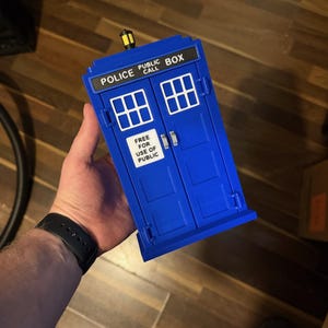 May include: A blue Police Box toy with white window details and the text "POLICE PUBLIC CALL BOX" and "FREE FOR USE OF PUBLIC". The toy has a yellow light on top.