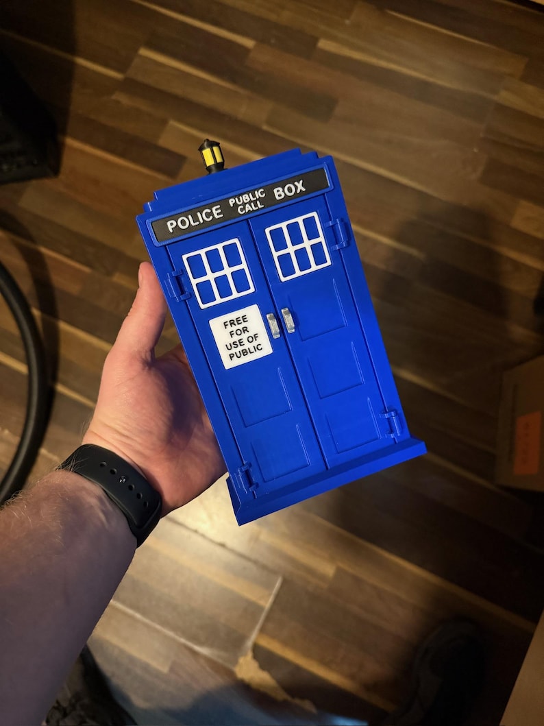 May include: A blue Police Box toy with white window details and the text "POLICE PUBLIC CALL BOX" and "FREE FOR USE OF PUBLIC". The toy has a yellow light on top.