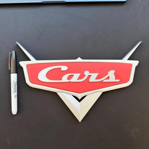 May include: A red and white "Cars" logo, with the word "Cars" in white script. The logo has a silver border and wings. A black Sharpie marker is next to the logo.