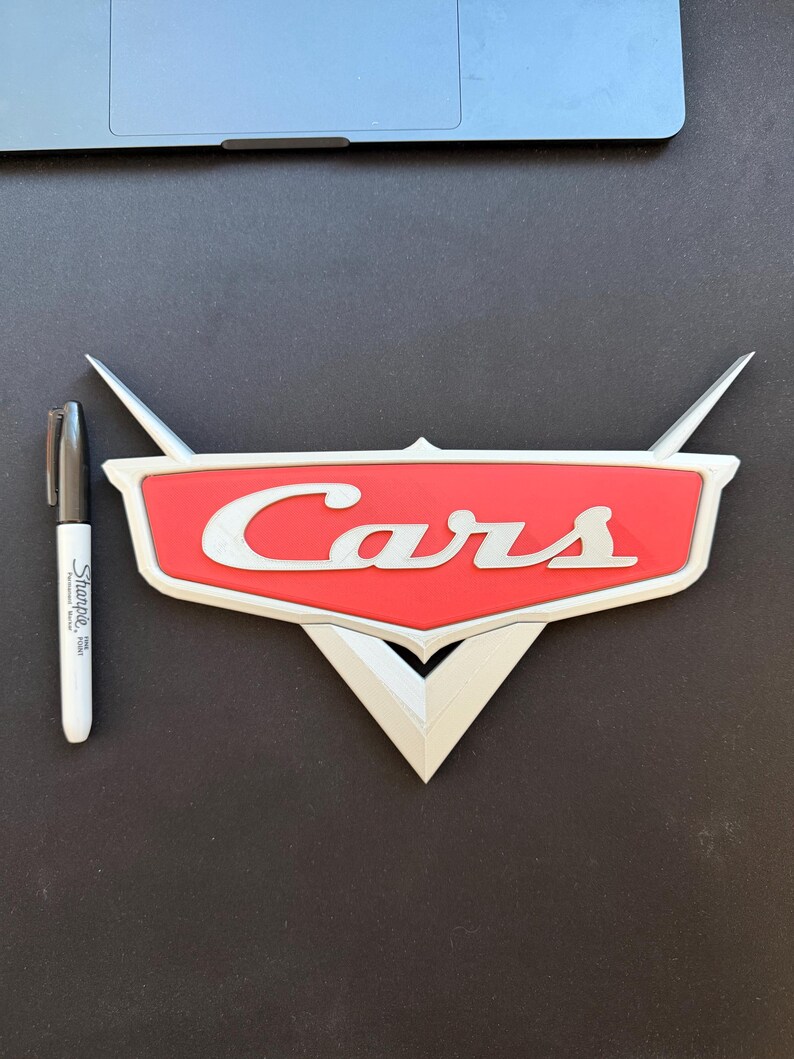 May include: A red and white "Cars" logo, with the word "Cars" in white script. The logo has a silver border and wings. A black Sharpie marker is next to the logo.