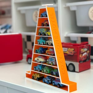 Op de afbeelding: Een oranje en witte displayplank in de vorm van een kegel, met de tekst "COZY CONE MOTEL" aan de zijkant. De plank bevat een verzameling miniatuur speelgoedauto's in verschillende kleuren. De plank staat op een wit oppervlak.