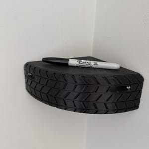 May include: A black, corner-mounted shelf with a tire tread design. A black Sharpie fine point marker rests on the shelf. The shelf is attached to a white wall with two screws.