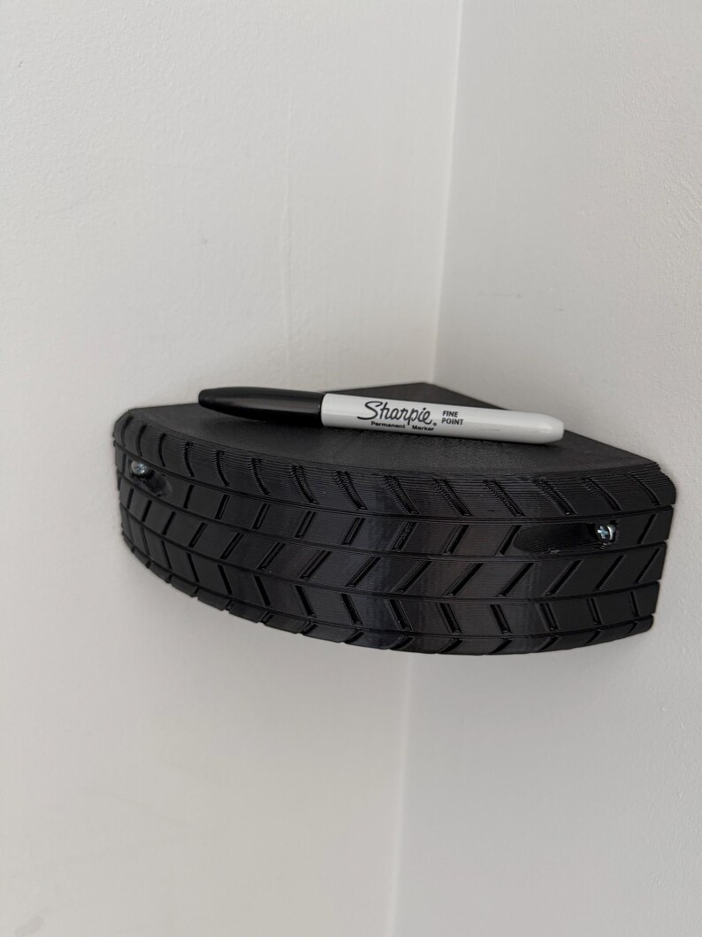 May include: A black, corner-mounted shelf with a tire tread design. A black Sharpie fine point marker rests on the shelf. The shelf is attached to a white wall with two screws.