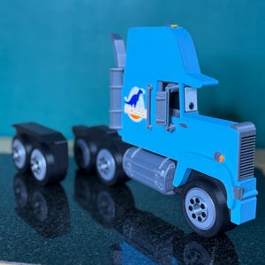 May include: A light blue toy semi-truck with a cartoonish design. The truck features a cab with a visor, a trailer, and a logo with a blue dinosaur. The wheels are gray and black.