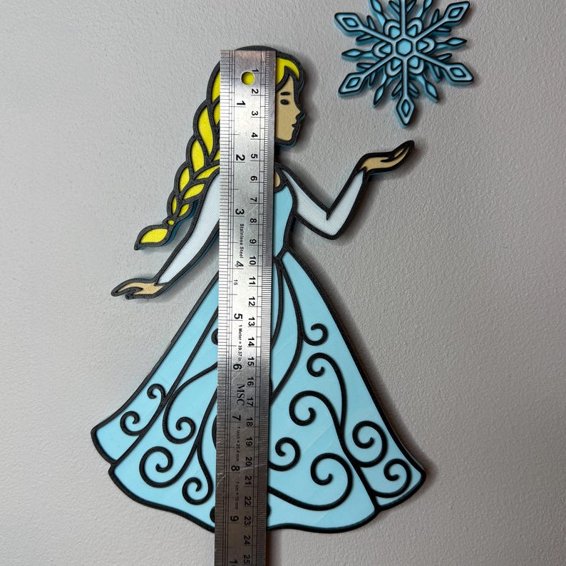 May include: A light blue and black cutout of a princess with a yellow braid and a snowflake. The princess is wearing a light blue dress with black swirls. A ruler shows the princess is approximately 9 inches tall.