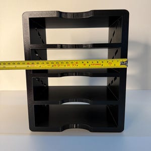May include: A black, three-tiered organizer with a rectangular shape. The organizer has a curved cutout on the top and bottom edges of each shelf. A yellow tape measure shows the width of the organizer is approximately 10 inches.