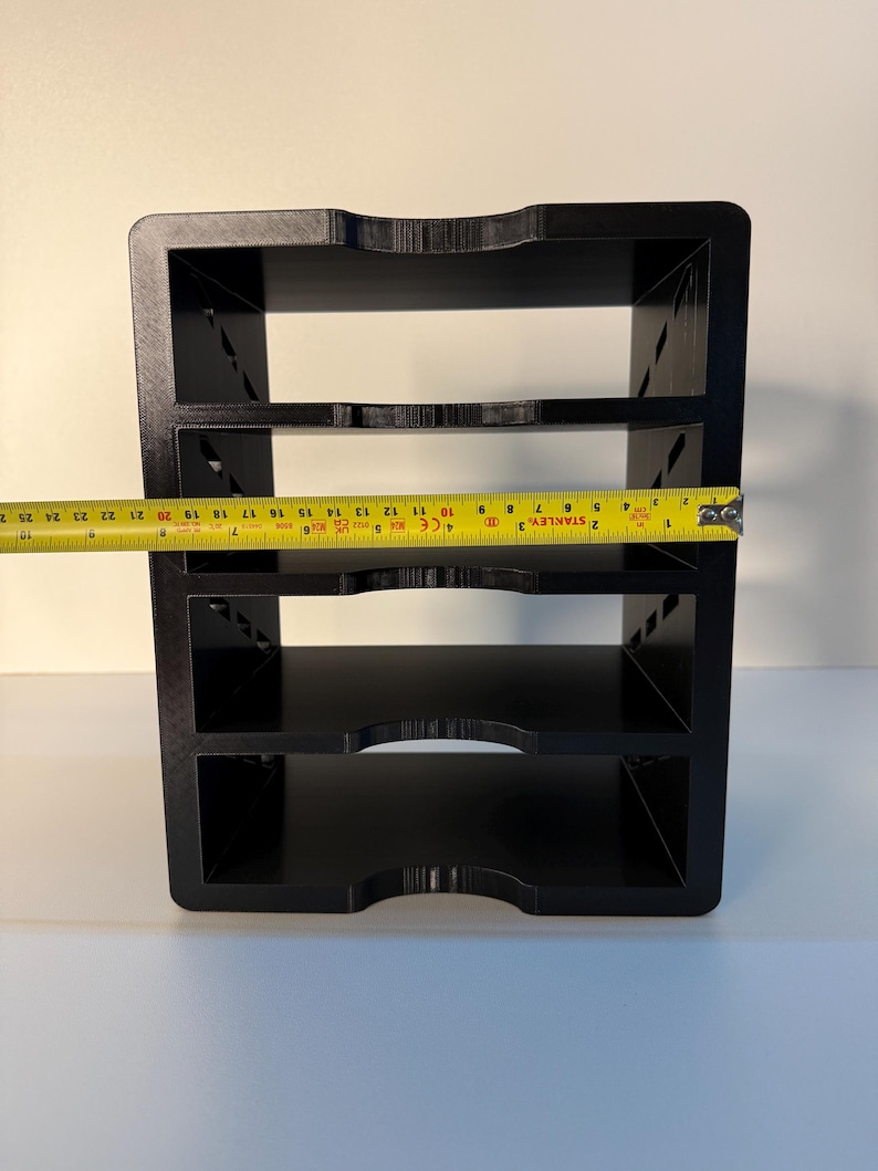 May include: A black, three-tiered organizer with a rectangular shape. The organizer has a curved cutout on the top and bottom edges of each shelf. A yellow tape measure shows the width of the organizer is approximately 10 inches.