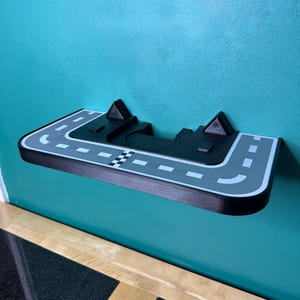 May include: A black and grey shelf designed as a race track, mounted on a teal wall. The shelf features a chequered flag, road markings, and two raised sections. The shelf is made of wood and has a curved edge.