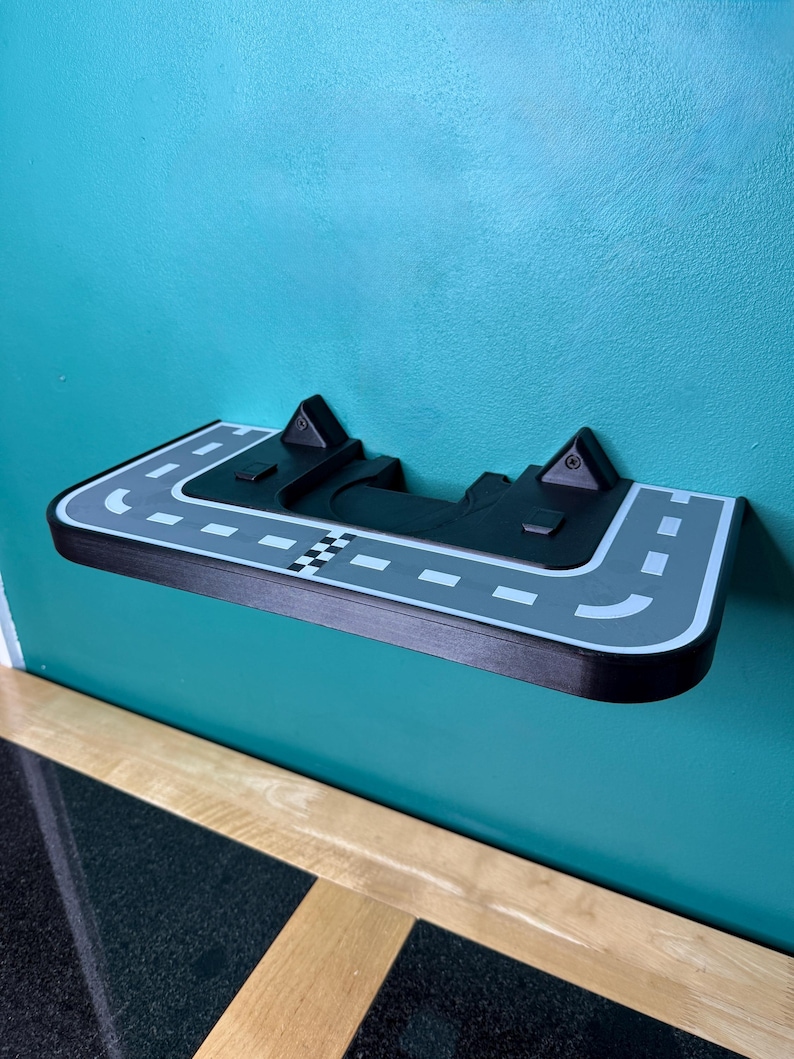 May include: A black and grey shelf designed as a race track, mounted on a teal wall. The shelf features a chequered flag, road markings, and two raised sections. The shelf is made of wood and has a curved edge.