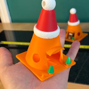 May include: An orange 3D-printed Christmas-themed decoration shaped like a traffic cone, topped with a red Santa hat and white trim. The cone has a small opening and a square base with two green Christmas trees. A second decoration is visible in the background.