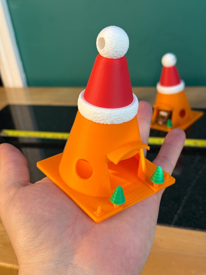 May include: An orange 3D-printed Christmas-themed decoration shaped like a traffic cone, topped with a red Santa hat and white trim. The cone has a small opening and a square base with two green Christmas trees. A second decoration is visible in the background.