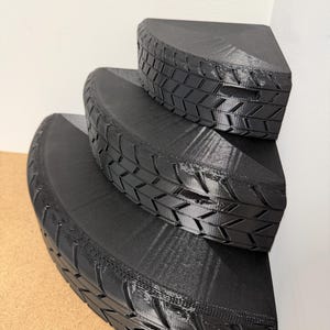 May include: Black, three-tiered corner shelf with a tire tread design. The shelves are semi-circular and fit snugly against a corner wall. The design gives the appearance of stacked tires, creating a unique storage solution.