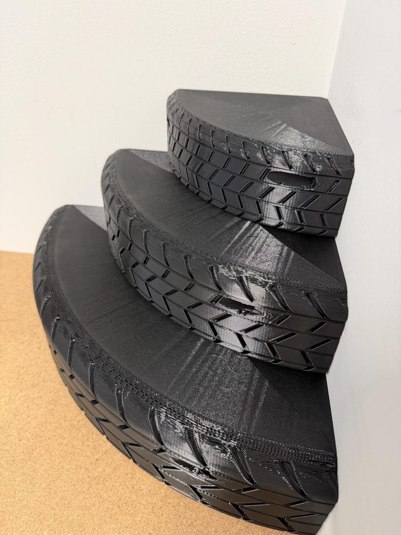 May include: Black, three-tiered corner shelf with a tire tread design. The shelves are semi-circular and fit snugly against a corner wall. The design gives the appearance of stacked tires, creating a unique storage solution.