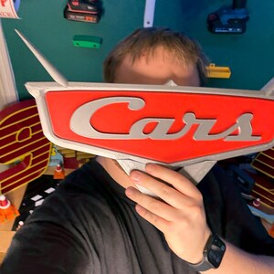 May include: A red and silver Cars logo, held in front of a person's face. The logo features the word "Cars" in silver lettering against a red background. The logo has silver wings extending from the top.