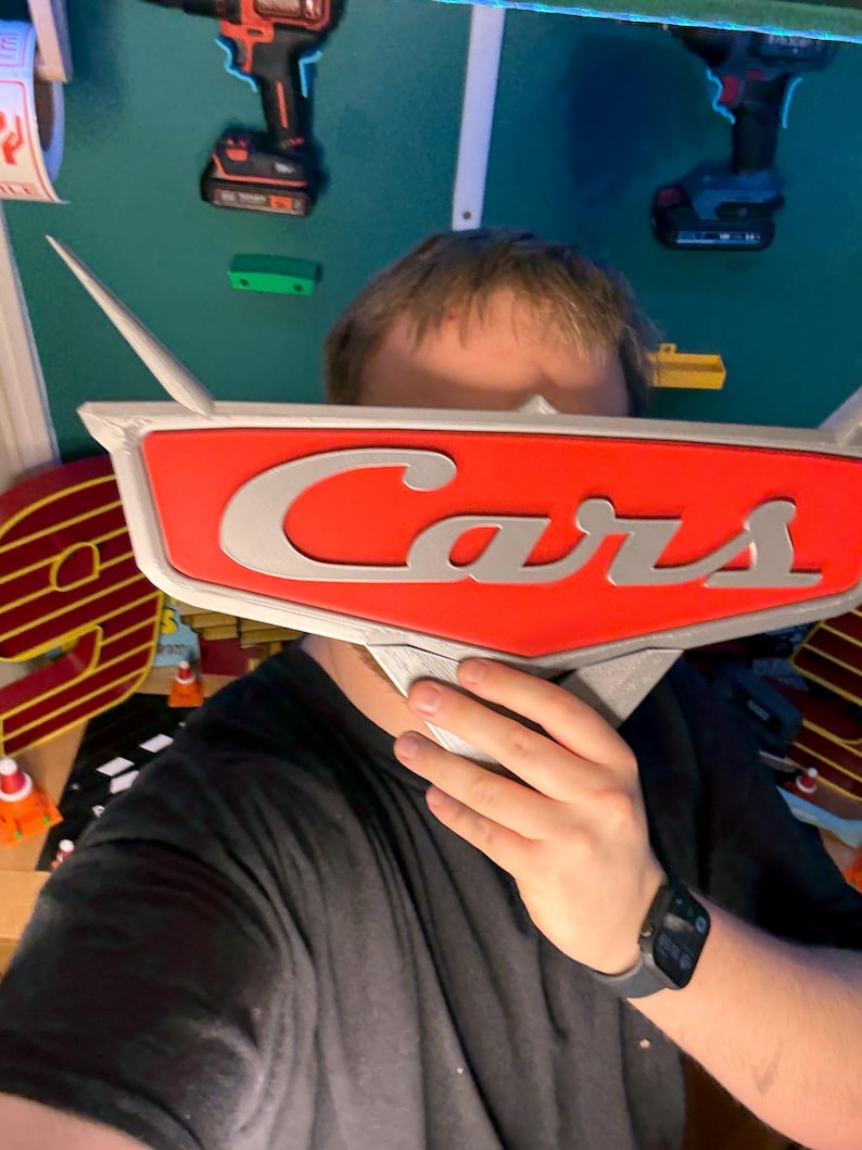 May include: A red and silver Cars logo, held in front of a person's face. The logo features the word "Cars" in silver lettering against a red background. The logo has silver wings extending from the top.