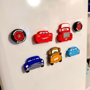 May include: A collection of colorful car-themed refrigerator magnets. The set includes magnets shaped like race cars, a truck, and tires. The magnets are red, blue, orange, and black, and are attached to a white refrigerator door.