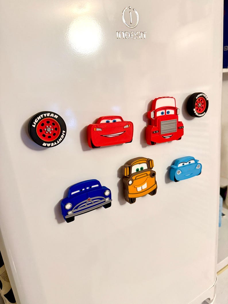 May include: A collection of colorful car-themed refrigerator magnets. The set includes magnets shaped like race cars, a truck, and tires. The magnets are red, blue, orange, and black, and are attached to a white refrigerator door.