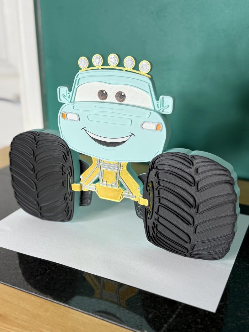 Op de afbeelding: Een lichtblauw monstertruckspeelgoed met grote zwarte banden en een geel chassis. De truck heeft een lachend gezicht en een rij gele lampjes bovenop. Het speelgoed is ontworpen om eruit te zien als een stripfiguur.