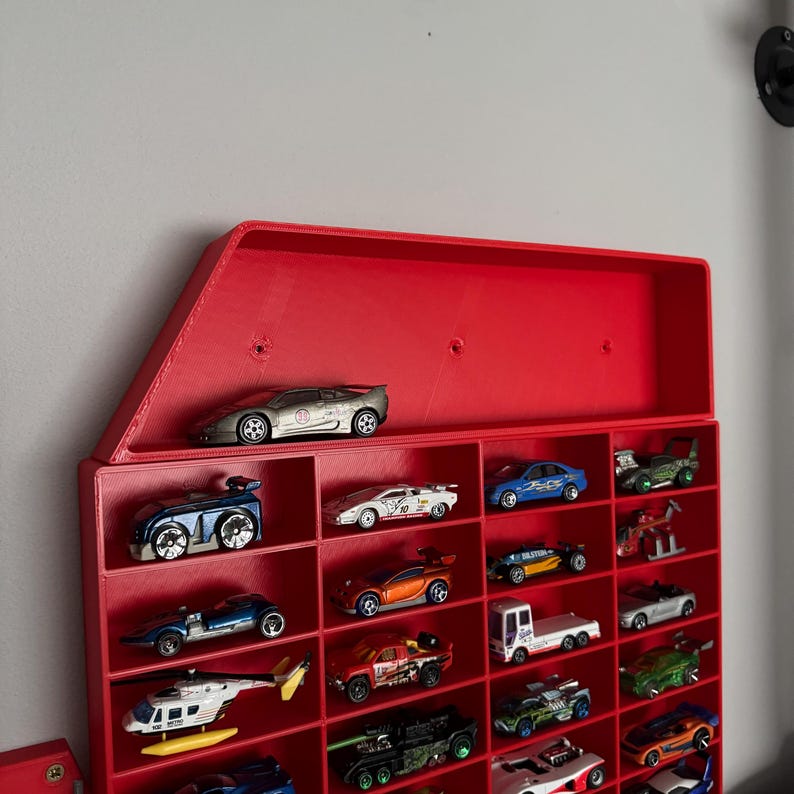 May include: A red plastic wall-mounted display case with multiple shelves for storing toy cars. The case is filled with various toy cars, including a silver sports car, a blue car, and a red truck.