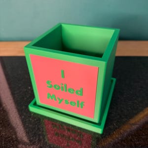 May include: A green square planter with a matching saucer. The planter has a red front panel with the words "I Soiled Myself" in green. The planter is sitting on a dark surface.