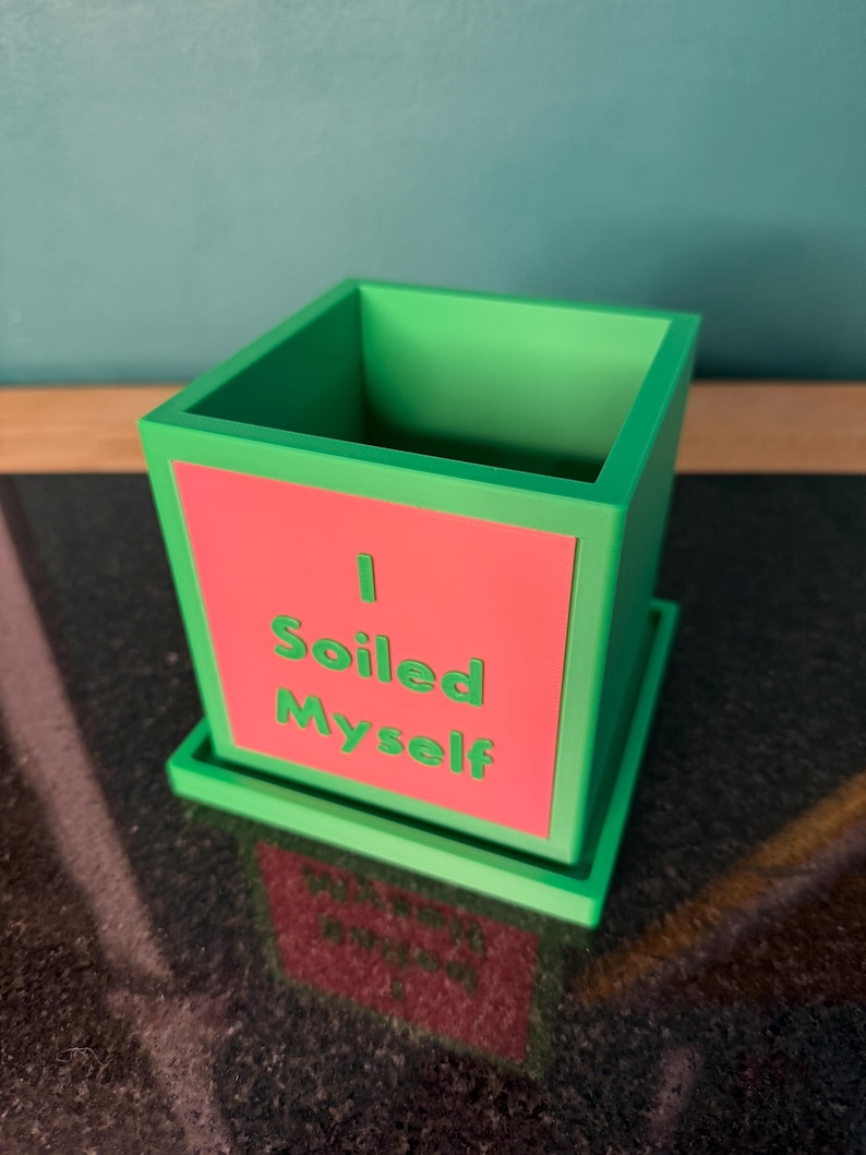 May include: A green square planter with a matching saucer. The planter has a red front panel with the words "I Soiled Myself" in green. The planter is sitting on a dark surface.