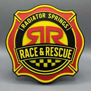 May include: A multi-layered, 3D-printed sign featuring the text "RADIATOR SPRINGS RACE & RESCUE". The sign is shaped like a fire department badge, with a red and yellow border, black accents, and a yellow center. The text is in black and red.