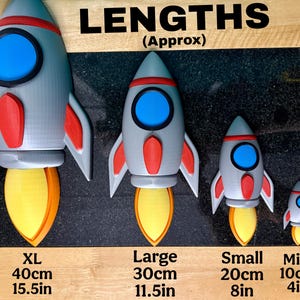 Puede incluir: Se muestran cuatro cohetes de juguete de diferentes tama&ntilde;os, de mini a XL. Los cohetes son grises con detalles en rojo y azul, y llamas amarillas y naranjas. El cohete XL mide 40 cm de alto, el grande 30 cm, el peque&ntilde;o 20 cm y el mini 10 cm.