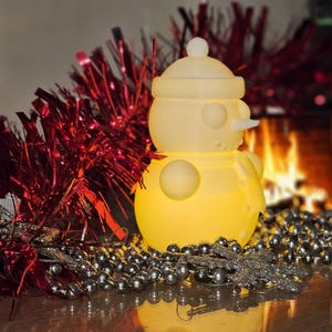 Yellow glowing 3D-printed snowman LED tealight holder, displayed on a festive table with red tinsel, silver beads, and a cosy fireplace in the background.