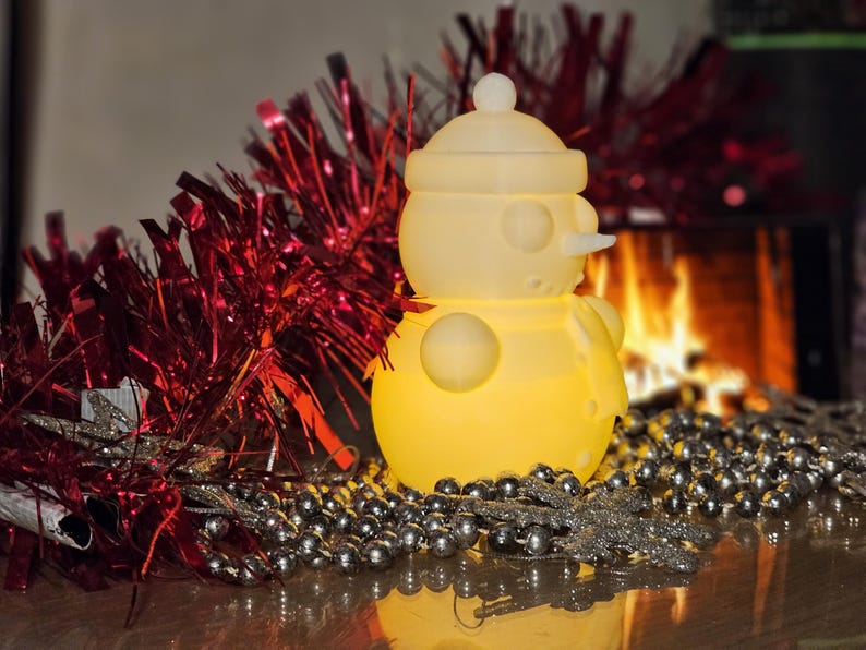 Yellow glowing 3D-printed snowman LED tealight holder, displayed on a festive table with red tinsel, silver beads, and a cosy fireplace in the background.