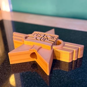 Puede incluir: Trofeo en forma de estrella naranja con el texto "Piston Cup" en el centro. El trofeo est&aacute; impreso en 3D y tiene un dise&ntilde;o en capas con un agujero en el centro de la estrella. Sobre una superficie oscura.