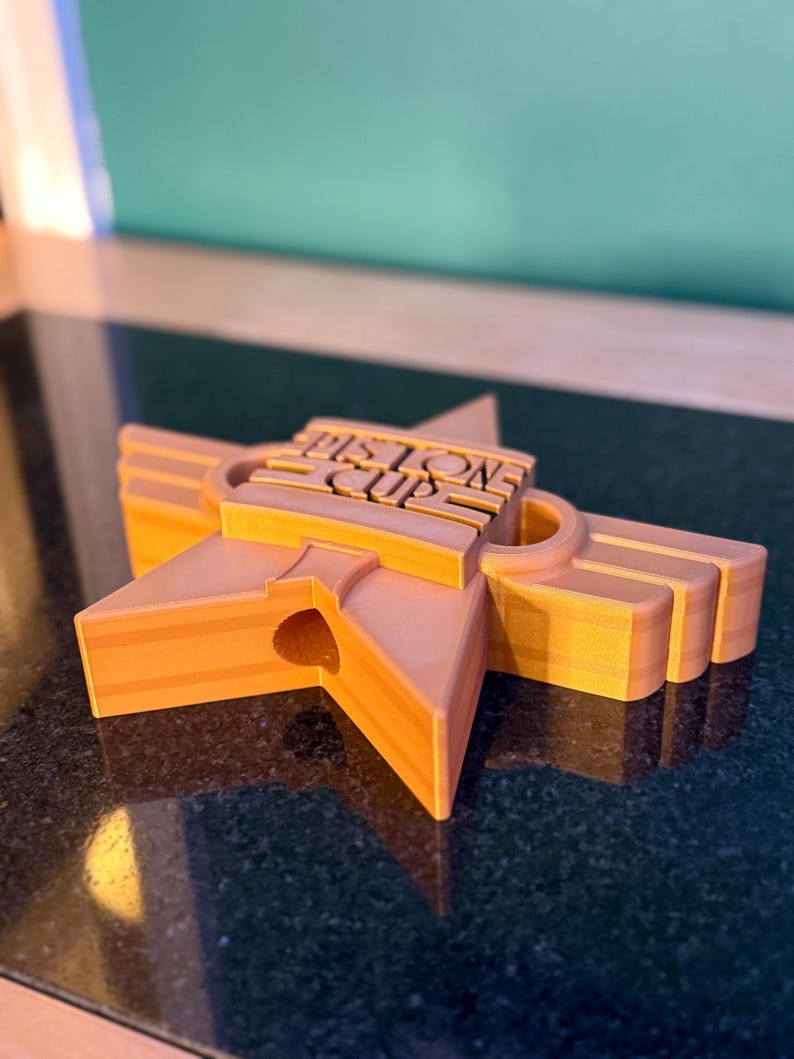 Puede incluir: Trofeo en forma de estrella naranja con el texto "Piston Cup" en el centro. El trofeo est&aacute; impreso en 3D y tiene un dise&ntilde;o en capas con un agujero en el centro de la estrella. Sobre una superficie oscura.