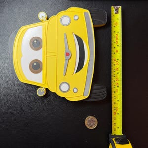 May include: A yellow cartoon car-shaped object with large eyes, black tires, and a red accent. The object is approximately 8 inches long, as measured by a yellow tape measure.