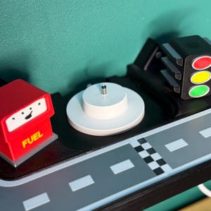 May include: A toy road scene featuring a red fuel pump, a white circular platform, a traffic light, and a grey road with white lane markings. The fuel pump has the word "FUEL" on it.