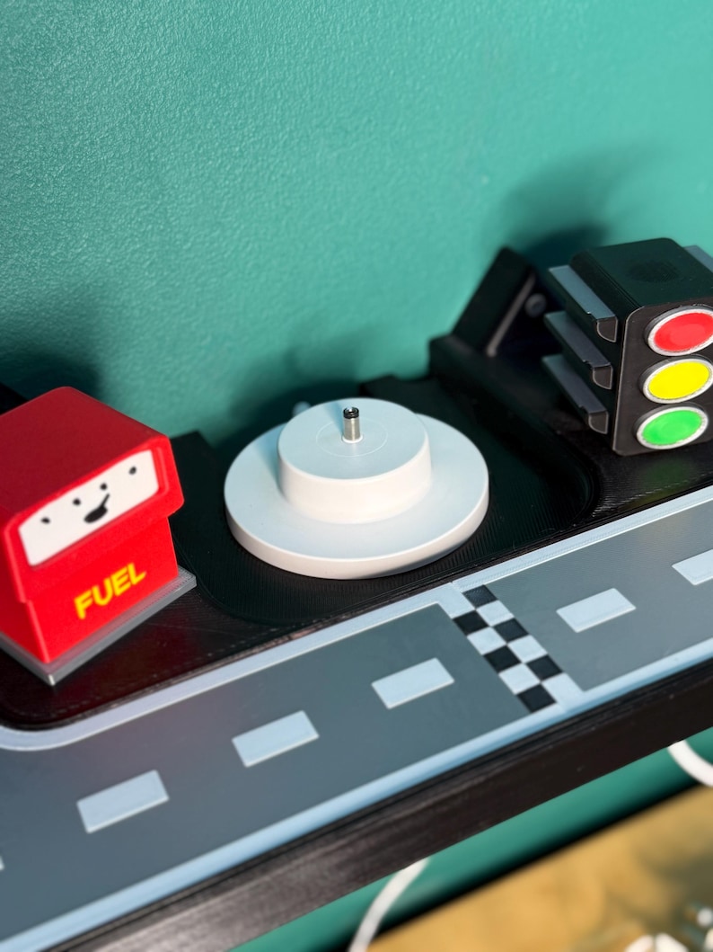 May include: A toy road scene featuring a red fuel pump, a white circular platform, a traffic light, and a grey road with white lane markings. The fuel pump has the word "FUEL" on it.