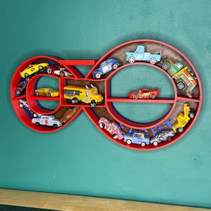 May include: A red infinity-shaped shelf filled with toy cars of various colors. The shelf is mounted on a teal wall. The toy cars include a yellow school bus, a red race car, and several other vehicles.