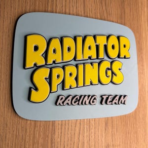 Puede incluir: Un letrero rectangular con esquinas redondeadas muestra las palabras "RADIATOR SPRINGS" en amarillo con contorno negro, sobre "RACING TEAM" en gris. El letrero tiene un fondo azul claro y est&aacute; montado sobre madera.