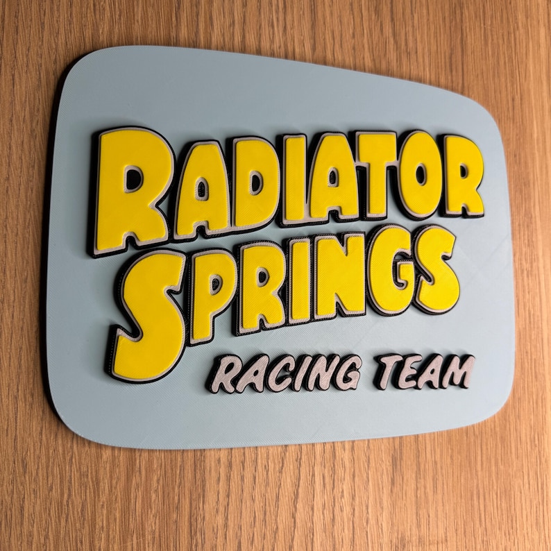 Puede incluir: Un letrero rectangular con esquinas redondeadas muestra las palabras "RADIATOR SPRINGS" en amarillo con contorno negro, sobre "RACING TEAM" en gris. El letrero tiene un fondo azul claro y est&aacute; montado sobre madera.