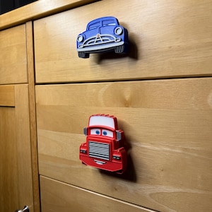 May include: Two car-shaped cabinet knobs. The top knob is blue, resembling a classic car. The bottom knob is red, resembling a truck. Both knobs have cartoon-like features and are mounted on a wooden cabinet.