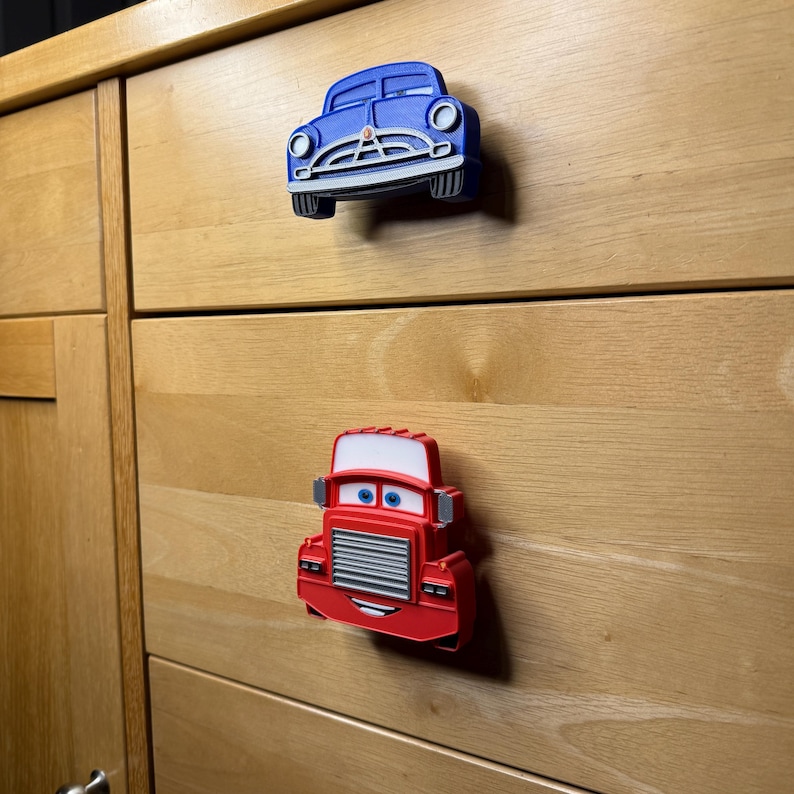 May include: Two car-shaped cabinet knobs. The top knob is blue, resembling a classic car. The bottom knob is red, resembling a truck. Both knobs have cartoon-like features and are mounted on a wooden cabinet.
