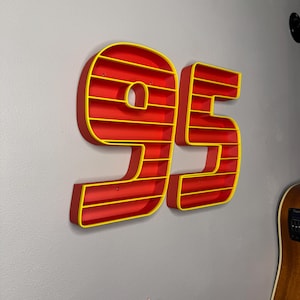May include: A wall-mounted shelf in the shape of the number 95. The shelves are red with yellow trim. The number is mounted on a gray wall.
