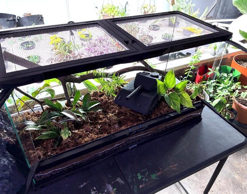May include: A rectangular glass terrarium with a black metal frame and a dual-hinged mesh lid. Inside, the enclosure features brown substrate, green leafy plants, a wooden branch, and a black toy tank model positioned among the foliage.